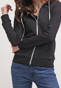 Sudadera negra con cremallera plateada, diseño ajustado, cordones blancos y puños acanalados. Combinada con vaqueros azules.