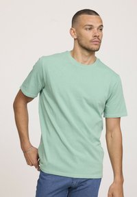 Homme portant un t-shirt à manches courtes vert clair et un jean bleu, regardant à droite avec la main près de la taille sur un fond uni.