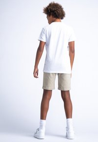 T-shirt bianca, pantaloni beige, sneakers bianchi. L'outfit presenta un design semplice e una vestibilità rilassata, mettendo in mostra linee pulite e colori neutri.