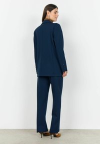 Blazer blu navy e pantaloni abbinati realizzati in un tessuto liscio. Il blazer presenta un colletto a taglio e una chiusura con un solo bottone, con polsini con bottone.