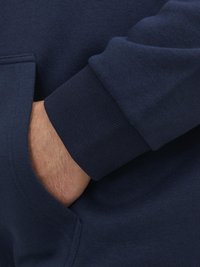 Marineblauwe hoodie met ribbels bij de manchetten en een voorzak. Zacht, gestructureerd materiaal zichtbaar op een close-up van een hand die in de zak rust.