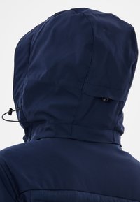 Marinblå vattentät jacka med avtagbar huva, som har dragskojustering och en slät tygtextur, utformad för utomhusbruk.