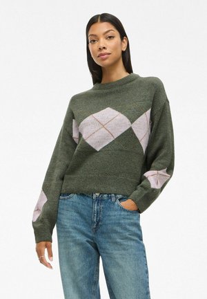 Femme aux longs cheveux noirs portant un pull en argyle vert et un jean bleu, posant avec une main dans la poche sur fond blanc.