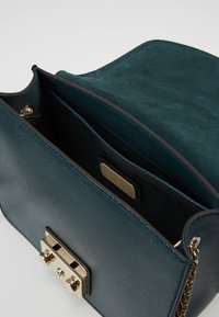 Borsa in pelle verde scuro con chiusura in oro e tracolla a catena, aperta per mostrare la patta foderata in suede e la tasca interna con etichetta del marchio.