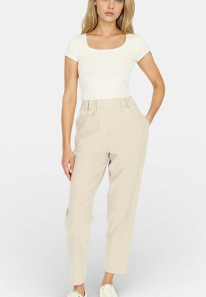 Kobieta ubrana w biały top z krótkim rękawem i dekoltem w kształcie łódki, beżowe spodnie z wysokim stanem i talią typu paperbag oraz białe trampki, stojąca z jedną ręką w kieszeni.