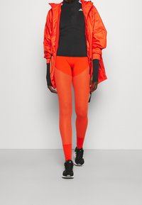 Osoba nosząca jasnopomarańczowe, teksturowane legginsy, pasującą pomarańczową kurtkę, czarną bluzkę z długim rękawem i czarne trampki Adidas, stojąca na jednolitym tle.