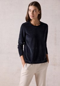Sweatshirt en polaire bleu marine avec manches longues, col rond et texture subtile. Associé à un pantalon beige clair, les mains dans les poches.
