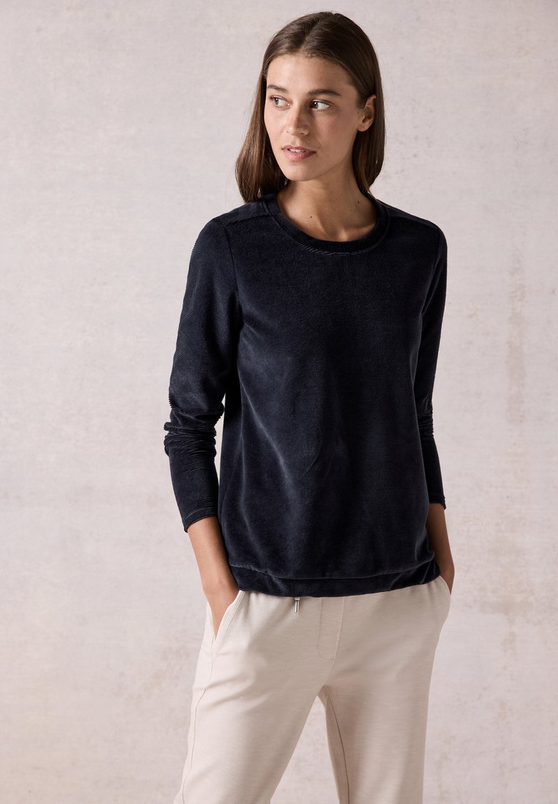 Sweatshirt en polaire bleu marine avec manches longues, col rond et texture subtile. Associé à un pantalon beige clair, les mains dans les poches.
