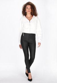 Hanorac alb crop cu fermoar în față, cu detalii ornamentale, asortat cu colanți negri din piele ecologică și pantofi negri cu toc ascuțit.