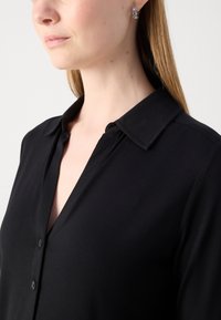 Blusa nera con colletto, chiusura a bottoni e texture morbida. Inquadratura ravvicinata che mette in evidenza il scollo e la finitura del tessuto liscia.