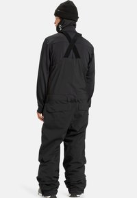 Man die met zijn rug naar de camera staat, in zwarte sneeuwbroek met bretels, een zwart longsleeve shirt, een zwarte beanie en winterlaarzen op een witte achtergrond.