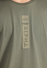 Olivno zelena majica s teksturirano gumo obloženim logotipom "ALPHA" i grafičkim logotipom u pravokutnom obliku na prednji strani.