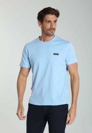 Homme portant un t-shirt bleu clair à manches courtes avec une poche avant et une étiquette "EST. 90", associé à un pantalon foncé, regardant sur le côté.