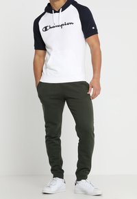 Młody mężczyzna w białej i granatowej bluzie krótkiego rękawa Champion, ciemnozielonych dresach i białych sneakersach, stojący z rękami w kieszeniach.