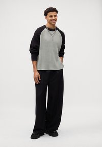 Grau gerippter Pullover mit schwarzen Ärmeln, lockere Passform; kombiniert mit lockeren schwarzen Hosen und klobigen schwarzen Schuhen; geschichtete Halskette.