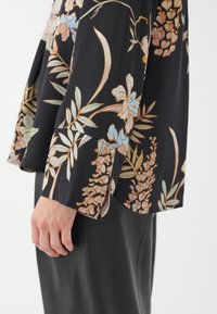 Schwarze Bluse aus Stoff mit floralem Design in hellblauen und pfirsichfarbenen Tönen. Mit lockeren Ärmeln und entspannter Passform. Akzente mit leichtem Glanz.