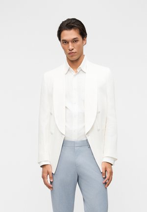 Bărbat purtând un sacou alb cu dublu rând de nasturi și o cămașă, cu pantaloni albastru deschis, stând în fața unui fundal simplu, deschis la culoare.