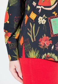 Camicia a stampa floreale in tessuto nero con colori vivaci, con varie piante e motivi di testo. Abbinata a pantaloni in velluto a coste rossi.