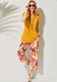 Haut jaune à manches courtes avec détails froncés ; pantalon blanc avec des motifs de feuilles colorées en rouge et orange. Le mannequin porte des lunettes de soleil et des baskets.