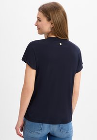 Navyblauwe t-shirt met korte mouwen en een ronde halslijn, zachte textuur en een klein metalen accent aan de achterkant. Gecombineerd met lichtblauwe jeans.