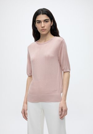 Femme aux longs cheveux foncés portant un haut en maille rose clair à manches courtes et un pantalon blanc, debout devant un fond blanc uni.