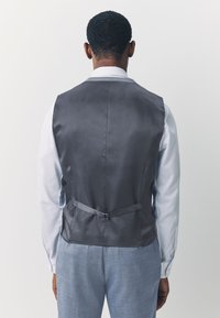 Gilet en satin gris à la texture lisse, avec une sangle ajustable à l'arrière, porté sur une chemise bleu clair, accompagné d'un pantalon fuselé en dessous.