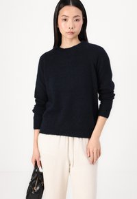 Navy blue knit sweater with a crew neckline, long sleeves, and a relaxed fit. Soft texture, paired with light-colored trousers and a black handbag.