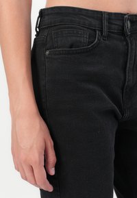 Svarta denimjeans med hög midja, framficka och en subtil textur. Närbild visar en hand som vilar mot tyget.