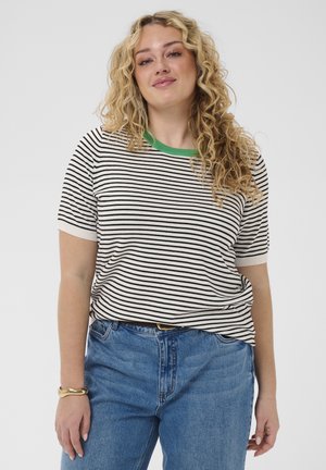 Hajlakkó, szőke göndör hajú nő, aki rövid ujjú fekete-fehér csíkos inget, zöld gallérral és kék farmert visel, egy egyszerű hátterű felület előtt áll.