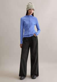 Hellblaues Langarmshirt mit hohem Kragen und gewelltem Saum, kombiniert mit weiten schwarzen Hosen. Das Modell trägt eine gestrickte graue Mütze.