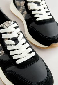 Melni sporta apavi ar teksturētu čūskas rakstu virsotni, baltām auklām un gumijas zoli. Materiāli ietver zamšādu un sintētisku audumu.