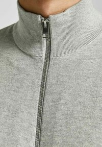 Šedý svetr na zip, vyrobený z měkké pletené látky. Má vysoký límec, kovový zip a žebrované lemy u výstřihu.