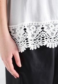 Top blanco con intrincado encaje negro en la parte inferior, combinado con pantalones de cuero negros. Primer plano de una mano reposando sobre la tela.
