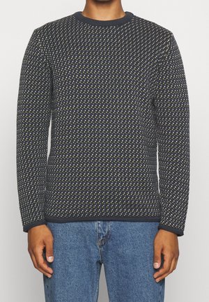 Mand iført mørkeblå sweater med små hvide og gule hjerte-lignende mønstre, kombineret med blå denimjeans, stående mod en hvid baggrund.