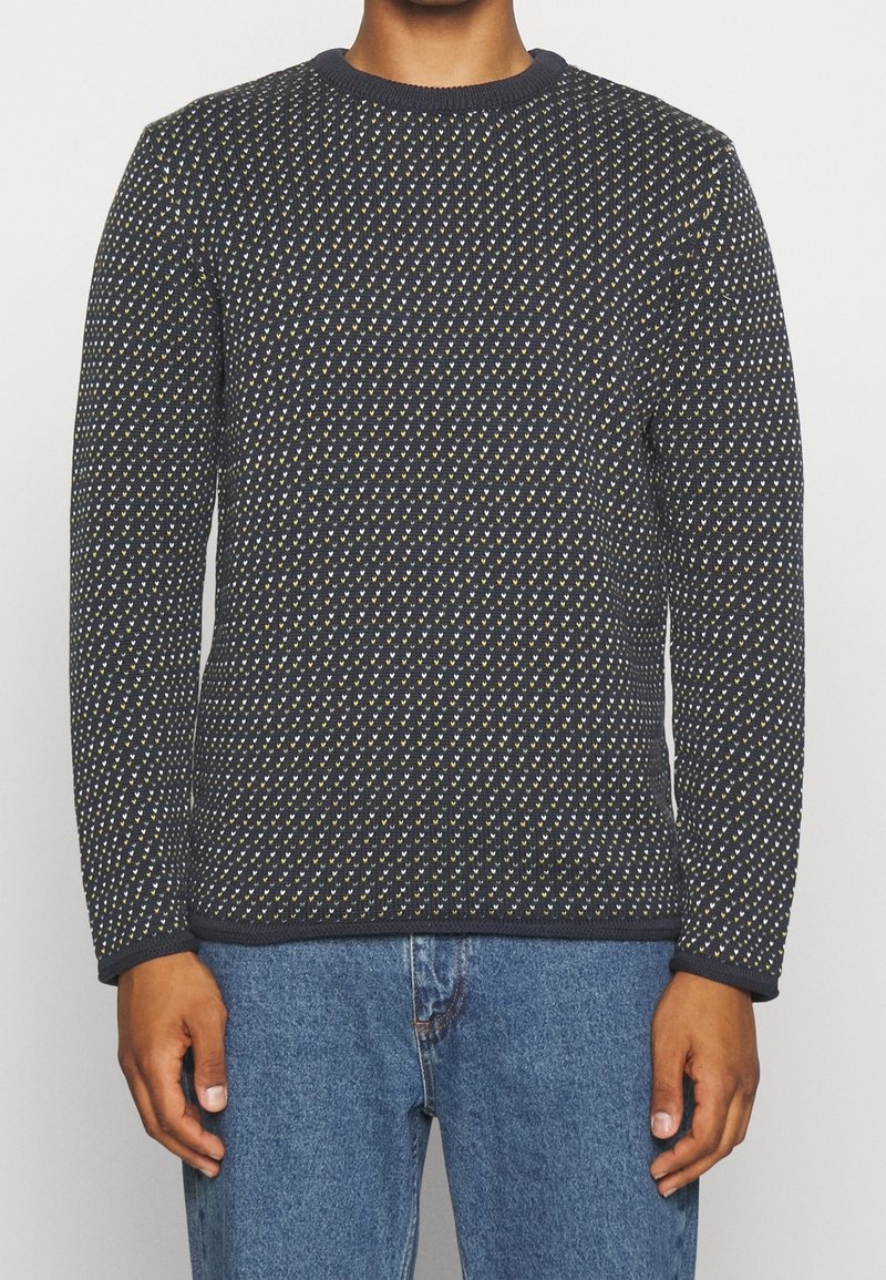 Homme portant un pull bleu marine foncé avec de petits motifs en forme de cœur blancs et jaunes, associé à un jean en denim bleu, debout devant un fond blanc.