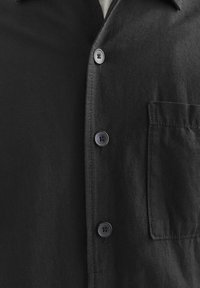 Chemise noire à boutons, en tissu lisse, avec un col pointu et une poche poitrine. Elle comporte cinq boutons foncés et une finition soignée.