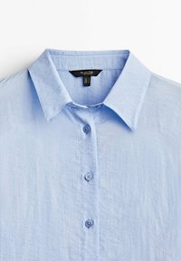 Jasnoniebieska koszula na guziki z ostrym kołnierzem, wykonana z lekko fakturowanej tkaniny. Z przodu znajduje się pięć guzików. Widoczna metka.