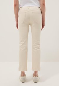Persona con pantalones rectos color crema y zapatillas blancas, de pie con los brazos relajados a los lados frente a un fondo sencillo.