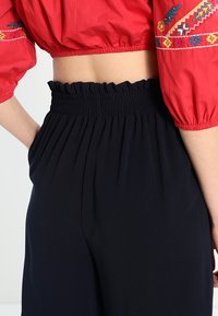 Röd blus med puffärmar pryds av färgglad broderi och har en kort design, i kombination med marinblå högmidjade byxor med elastisk smock.