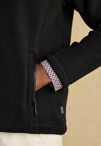 Persona che indossa una giacca in pile nera con un polsino a quadri visibile, mano nella tasca della giacca, sfondo beige.