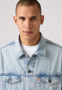 Mladenič s kratkimi lasmi, oblečen v svetloblu di denim jakno preko bele srajce, gleda neposredno v kamero na enotnem ozadju.