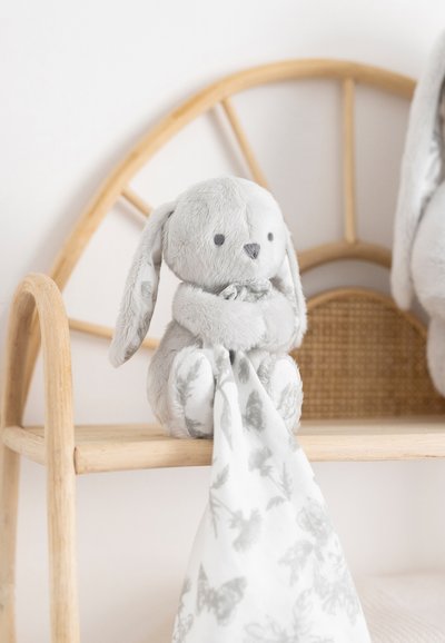 Pehme hall jänes pika kõrvadega, hoidmas valget kangast teki hallide liblikate mustriga, istub puidust riiulil.