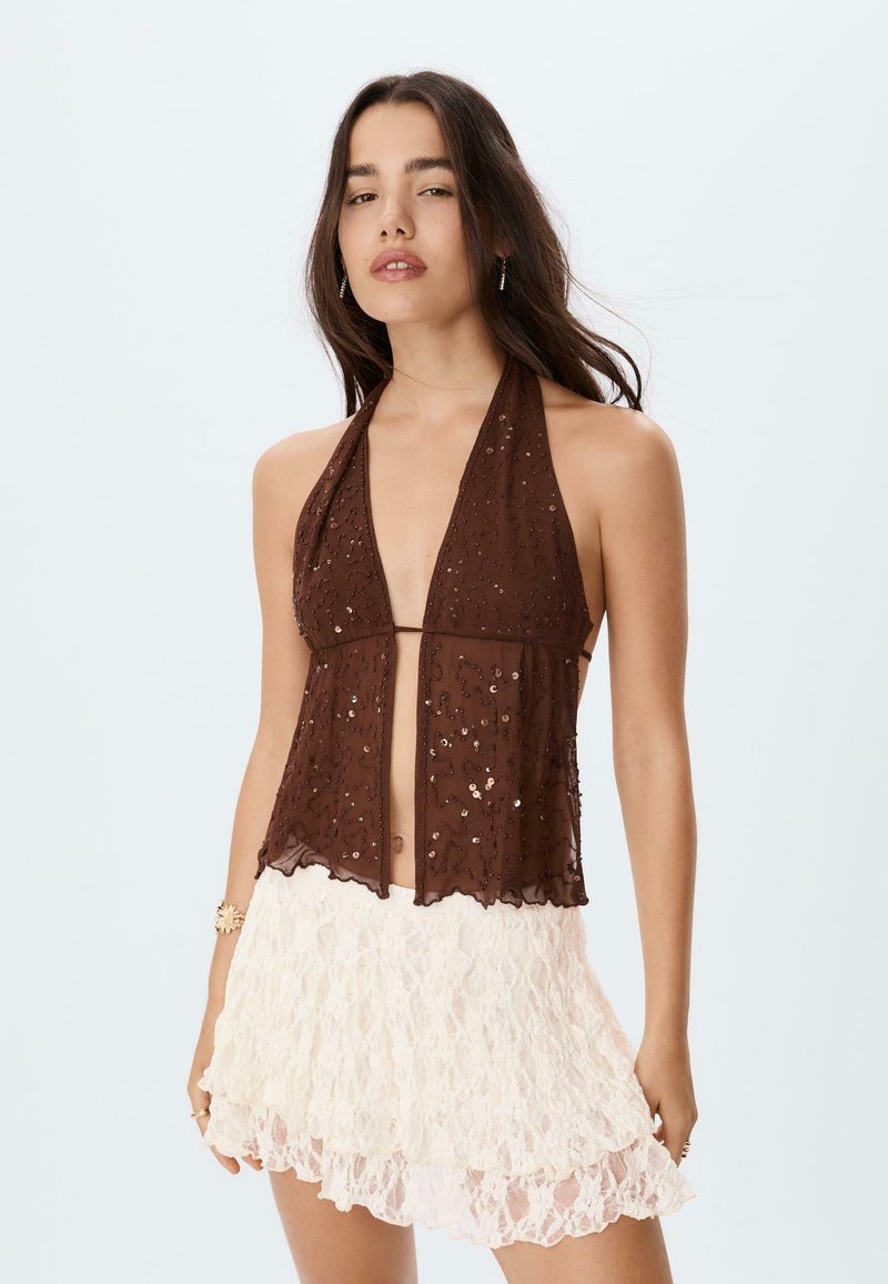 Hnědý halter top s průhlednou látkou a detaily z flitrů, s hlubokým výstřihem do V a otevřeným předním dílem. Vytvořeno v kombinaci s bílou krajkovou mini sukní.