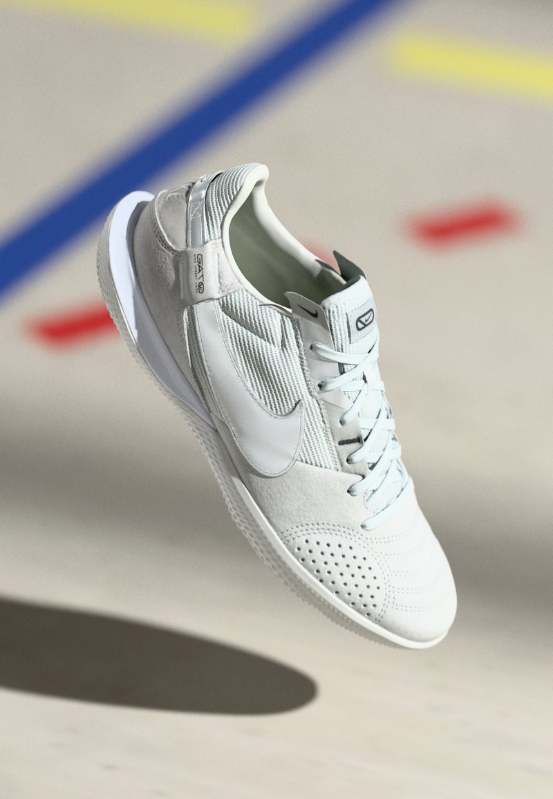Nike sneaker világosszürke színben, velúr és hálós textúrákkal, perforált részletekkel, fehér talppal és ezüst akcentussal a sarkán.