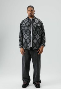 Man met ingevlochten haar, gekleed in een grijs-zwarte jas met diamantpatroon, zwarte wijde broek en zwarte schoenen, staand tegen een effen lichte achtergrond.