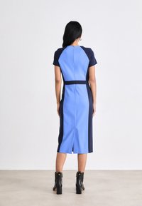 Φόρεμα εφαρμοστό με σχέδιο μπλοκ χρωμάτων μπλε και ναυτικό, κοντά μανίκια, φερμουάρ πίσω και σκίσιμο μέχρι το γόνατο στο στρίφωμα. Μαύρες μπότες μέχρι τον αστράγαλο.