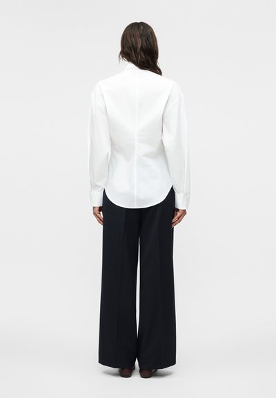 Γυναίκα με σκούρα μαλλιά, φορώντας εφαρμοστό λευκό μακρυμάνικο πουκάμισο και μαύρο, φαρδύ παντελόνι, όρθια με την πλάτη προς τα εμπρός σε απλό φόντο.