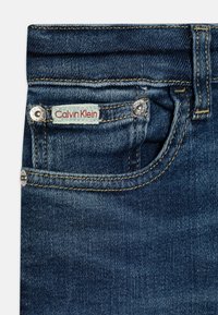 Sötétkék farmernadrág elülső zsebbel, ezüst hardware-rel és piros, világoskék Calvin Klein logo címkével. Látható varrási részletek.