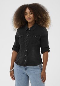 Schwarzes Denim-Hemd mit Kragen, hochgerollten Ärmeln, Knopfleiste und zwei vorderen Taschen. Kombiniert mit hellen Jeans.