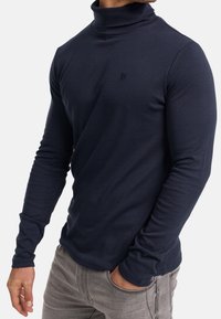 Homme portant un pull à col roulé bleu marine ajusté à manches longues et un jean gris clair, une main dans la poche.